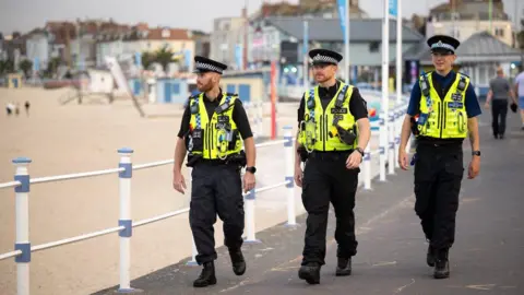 Three police officers walking along a beach area on a cloudy day. Some people behind them are walking away, others can be seen on the sand.