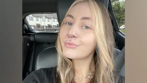 A selfie Kelly has taken in a car with black leather seats. Her seatbelt is covered by long, wavy brunette hair which has blonde dye through it. She is wearing a black jumper with a striped pattern. A silver necklace made of a string of heart shapes rests on her neck. Kelly, who has green eyes, is pouting and has multiple piercings, including a silver stud in the centre of her upper lip. She has two silver hoops on her outer nostrils and a septum piercing, which has two pyramid shaped ends, one coloured pink and the other blue.  