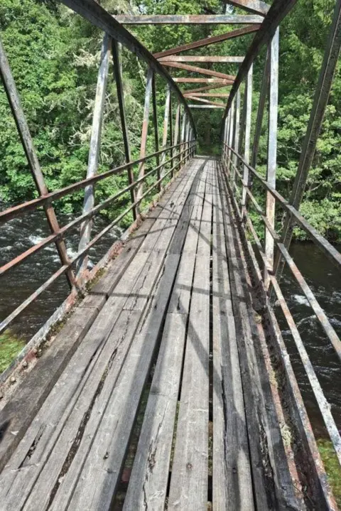 Forestry and Land Scotland This image is taken looking along the top of the bridge from one side to the other. The decking boards look in poor condition. The metal work structure of the bridge has rust coloured patches. 