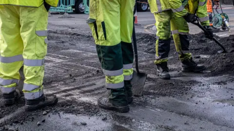 Close-up shot of the legs of three workers in hi-vis overalls working on a dark-grey road surface. All three are wearing black boots and are holding spades. The one on the right is shovelling black soil.