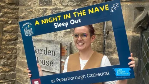 Light Project Peterborough A woman wearing a white shirt and a brown waistcoat holding a blue cut-out promoting the event. She is smiling at the camera.