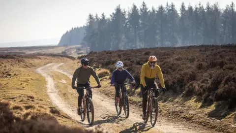 Mick Kirkman Three cyclists riding on a gravel route with some trees in the background. Two of the cyclists are men, one is wearing a green top and a black helmet, the other is wearing a mustard hoodie and an orange helmet. The other cyclist is a woman and is wearing a blue hoodie and a grey helmet.