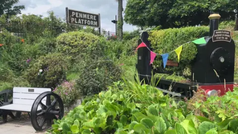 A minature train covered in greenery and glags, it says Penmere Platform Centenary of Opening 1st July 2025 on it. There is also a sign which says Penmere Platform. 