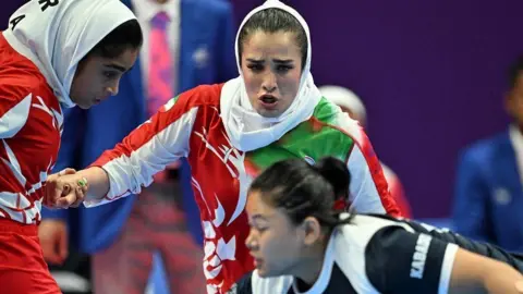 Getty Images Two women, wearing white head scarfs and red and white kits, are focused on another woman, who is wearing a black and white kit. 