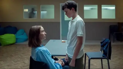 Netflix A woman is sitting on a chair looking up at a teenage boy who stands too close and looks down at her in a threatening way. They are alone inside a room with nothing but a table and two chairs, and three green and blue beanbags in the back of the room.