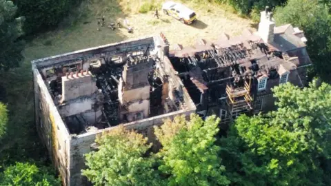Chris Bainbridge A drone image of Armathwaite Castle showing the damage caused by a fire. The building has a main rectangular structure with no roof and another structure connected to it, which is more similar to a house in shape. The roof is partially still on this section. Inside the affected areas are completely black with debris and collapsed beams and supporting structures. There is a lawn with an emergency vehicle parked on it and a group of people standing nearby.