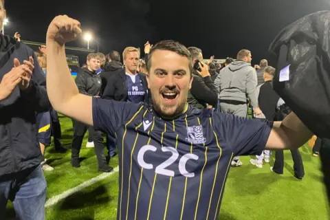 Warren Muggleton/BBC A man wearing a dark blue Southend United jersey is raising his fists cheering in front of the camera. Behind him are other young men crowding on the pitch at Forest Green Rovers.