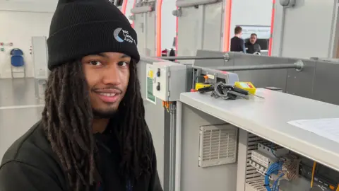 e Louis Barklem is wearing a black beanie hat and a black top.  He has a short trimmed beard and dreadlocks.  He is looking at the camera as he is working on wiring in a metal cabinet in a grey-coloured room. Two people are visible through a gap behind the cabinet.