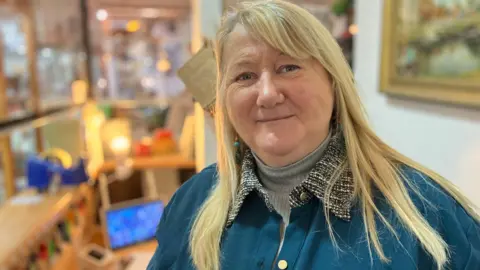 A woman with blonde hair wearing a blue jacket looks into the camera.