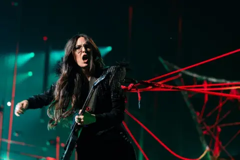 EBU / Corinne Cumming Veronica Fusaro plays guitar while straining against a web of red ropes