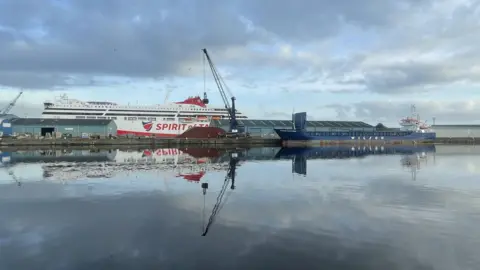 The Spirit of Tasmania berthed in Leith. The ship is partially obscured by buildings. A barge sits in the water in the foreground. It is blue. The other ship is mainly white, with a red hull and the words 'Spirit of Tasmania' in red on the side.