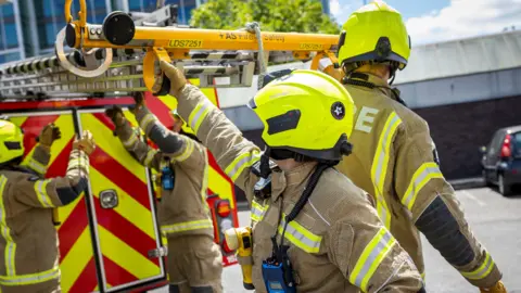 Four firemen standing by a fire truck pulling a large piece of equipment off the roof of the vehicle. They are all in their uniform with a yellow hi vis helmet on. 