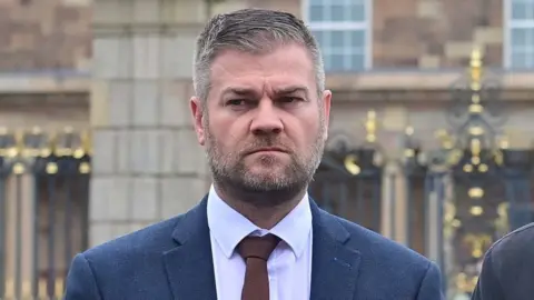 Colin McGrath with greying hairsbreadth  and a beard stares to his close    with a stern look   connected  his face. He is wearing a blue/grey suit, achromatic  necktie  and brownish  tie.