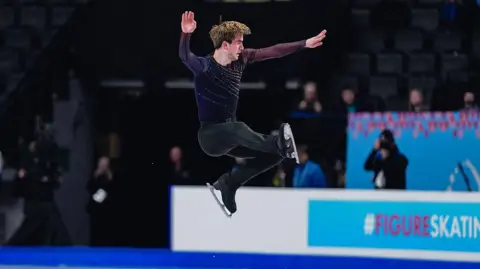 Getty Images Edward Appleby competing on the ice. He is mid-jump and fully in the air. The crowd is blurred in the background. 
