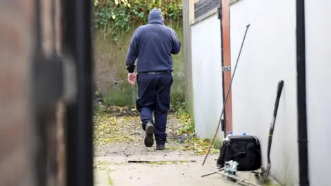 Niall Carson/PA Wire A member of the police team involved in the search for Kieran Durnin. The man is wearing dark blue clothes, boots and a beanie hat. He walks down the alley towards a wall covered in ivy. In the foreground are a garden fork, a bag and bottles.