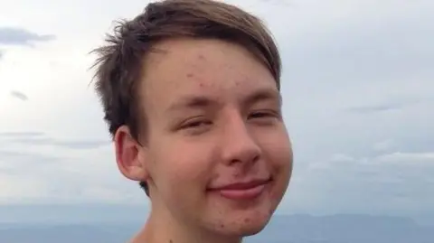 British Transport Police Sam, a white man, with short brown hair, smiles at the camera. He appears to be beside a body of water and some mountains