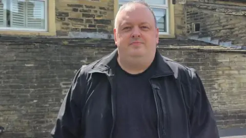 Ian Hudson Man starting directly at the camera. he is wearing a dark coat and a dark top. Behind him is an older style building and brickwork.