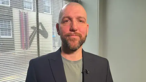 BBC News Deejay Whittingham standing in front of a window and blinds looking into the camera. He is wearing a t-shirt and blazer. He has a beard and bald head.
