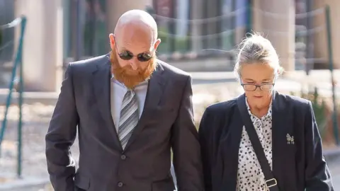 Athena Picture Agency Neil Lewis wearing a suit and walking along with a blonde woman. There heads are both down. 