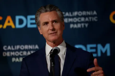 Getty Images Gavin Newsom wearing a blue suit and white shirt, is speaking into a microphone at a podium in front of a poster for Prop50, his redistricting ballot measure which was passed by voters this week