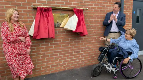 Paralympic gold medallist Tanni Grey-Thompson, who uses a wheelchair, pulling a golden cord to open a set of two red curtains. They reveal a golden plaque to mark the reopening of the station. Grey-Thompson smiles and has short, blonde hair, glasses, and is wearing a light blue top with white polka dots. She is joined by Councillor Lisa Evans, leader of Stockton Council, and Tees Valley Mayor Ben Houchen.