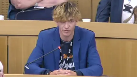 City of Doncaster Council A man sitting at a wooden bench in a blue suit jacket. He has bleached hair and a black t-shirt, speaking into a microphone.