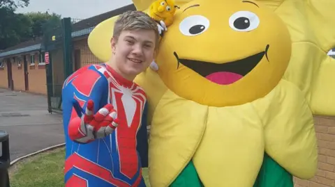 A teenage boy with short blonde hair is wearing a spiderman costume and standing next to a giant yellow cartoon flower 