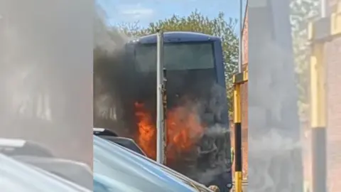 The back of a blue bus. The picture has been taken from behind the vehicle and flames can be seen coming from it. 