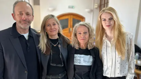 A man and three women inside a building. The three on the left are wearing black and the woman on the right is in white