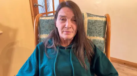 Carla Fowler/BBC A woman sat down on a high backed chair looking straight ahead. She has long, dark brown hair and is wearing a teal coloured hooded jumper. 