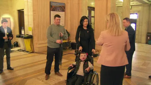 A photo from the event at Stormont showing Michael McMonagle standing to the left of the screen looking down at his phone, he is wearing trousers, shirt and a jacket. Michelle O'Neill stands to the right of the screen with her back to the camera, wearing a pink jacket. In the centre of the image are Daíthí Mac Gabhann in a wheelchair and his parents standing behind him.