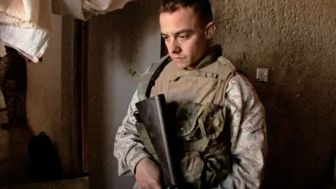 Michael Epstein A man in camouflage clothing with a khaki vest over the top holds a gun and looks down at the ground. Behind him is a rough beige wall. 