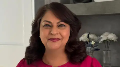 Sbba Siddique is smiling at the camera and has shoulder length dark hair. She is wearing a pink top and there are two vases of flowers behind her. 