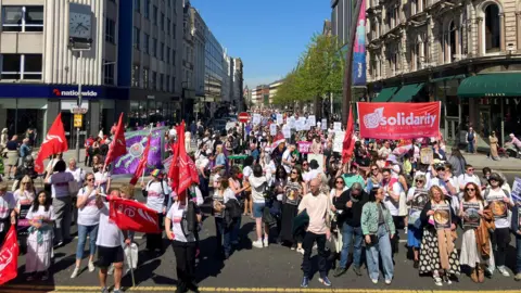 A large crowd are standing on a road. Many are holding banners that says solidarity. Some people are holding photos of women. Shops in a high street are in the background. 