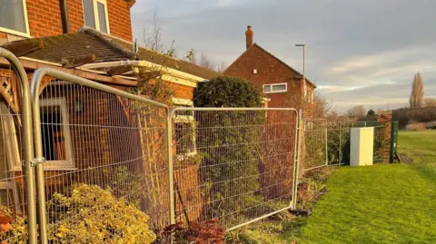 Mark Palmer Heras fencing installed on the very edge of residents' homes. Their back gardens are on the left of the image, inches away from the fencing which blocks the view from their windows. On the right side of the fencing is a lush green field. 