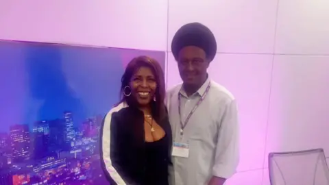 BBC presenter Merisha Steveson with Mykal Brown standing next to a backdrop of the Birmingham skyline at night in a room with purple lighting