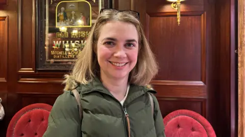 Georgia Hussey pictured in Harp Bar Belfast, wearing a green puffer jacket - she has shoulder length blonde hair, there is wood panelling on the walls, an Old Irish Whiskey mirror and red velvet seats