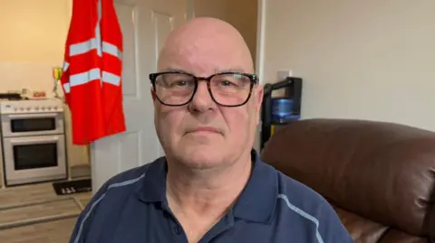 The photo shows a man smiling at the camera. He has brown rimmed glasses and is wearing a navy polo top. He's sitting on a brown leather sofa. Behind him is a door which is open, and has a bright orange coat hung from it. There is a white oven behind that. 