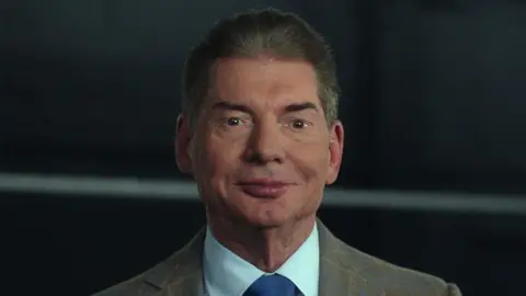 A still from Netflix show Mr.McMahon in which Vince McMahon, a man wearing a brown blazer, white shirt and blue tie, with white ring ropes in the background.