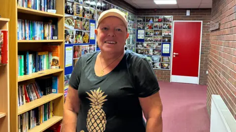 LDRS A woman with a black graphic t-shirt with a picture of a pineapple on it. She is wearing a cap and smiling.