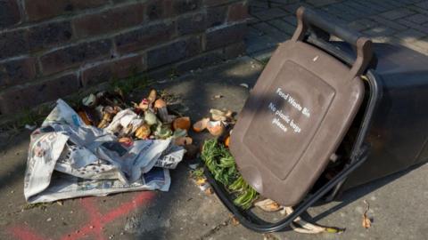 A brown lidded food waste bin lies on its side on the pavement with food waste spilling out 