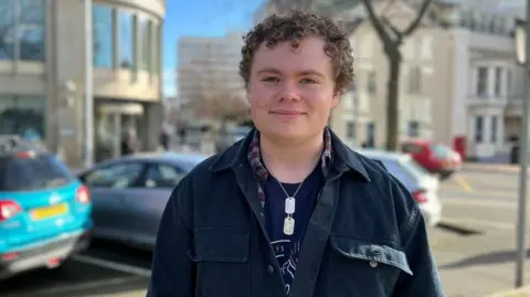 A young man with medium-length, brown, curly hair looks into the camera lens. He is wearing a navy jacket over and red and white checked shirt and blue t-shirt. He is wearing a necklace with two silver pendants.