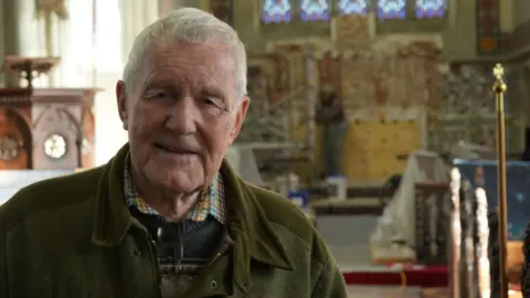 Jamie Niblock/BBC Warden Christopher Francis stands in the church with his back to the altar where the restoration work is taking place. he wears a shirt, jumper and dark green jacket. The lectern can also be seen behind him.