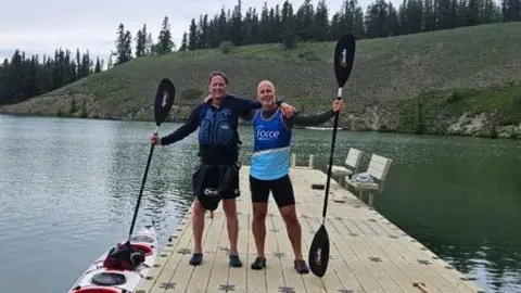 Jim Nye Ian Tyers and Jim Nye are standing on a jetty wearing shorts, long sleeved black T-shirts and holding black paddles. They have a two person kayak moored to the jetty to the left of the picture. Jim is wearing a blue vest with the word Force on it.