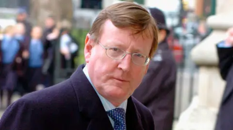 Getty Images Mr Trimble is looking into the camera. He's wearing glasses and has brown hair. His coat is navy and he's wearing a blue patterned tie and a blue stripped shirt.