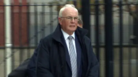 Frank Daly is an older man with short white hair. He is wearing a pair of rectangle glasses, a navy coat, a white shirt and a striped tie. The background of the courthouse is blurred.