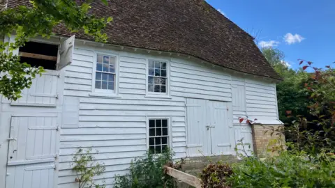 JOANNA TAYLOR/BBC The exterior of the watermill. It is a large building made from white wooden panels, with a brown brick roof. 