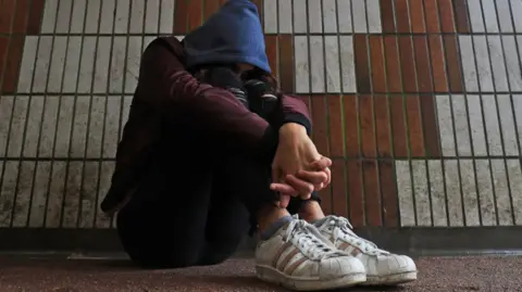 The picture shows a person sitting on the ground in what looks like an underpass or tiled walkway. Their head is covered by a hood, and they are curled up with their arms wrapped around their knees. Their face is not visible. The person appears to be wearing trainers, dark trousers and a jacket.