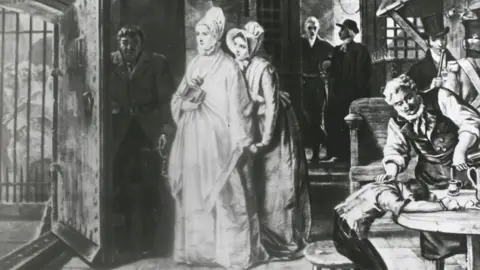 Getty Images A black and white drawing of Elizabeth Fry in Newgate Prison in the early 19th century. She approaches a wooden prison door on the right, which is opened by a locksmith with a large key. Through the door you can see bars and behind them the faint figures of female prisoners. Behind her is another woman and behind her two men standing at a door. On the far right, a boy and a man sit at a wooden table.