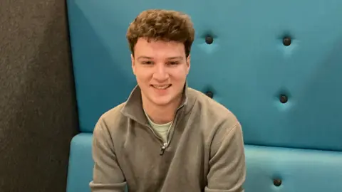 A young man with brown curly hair and a grey fleece on blue-backed chairs